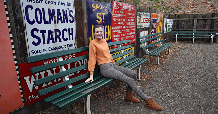 disabled blogger Kate Stanforth sat on bench in Beamish Museum with collection of historic signs on wall behind her